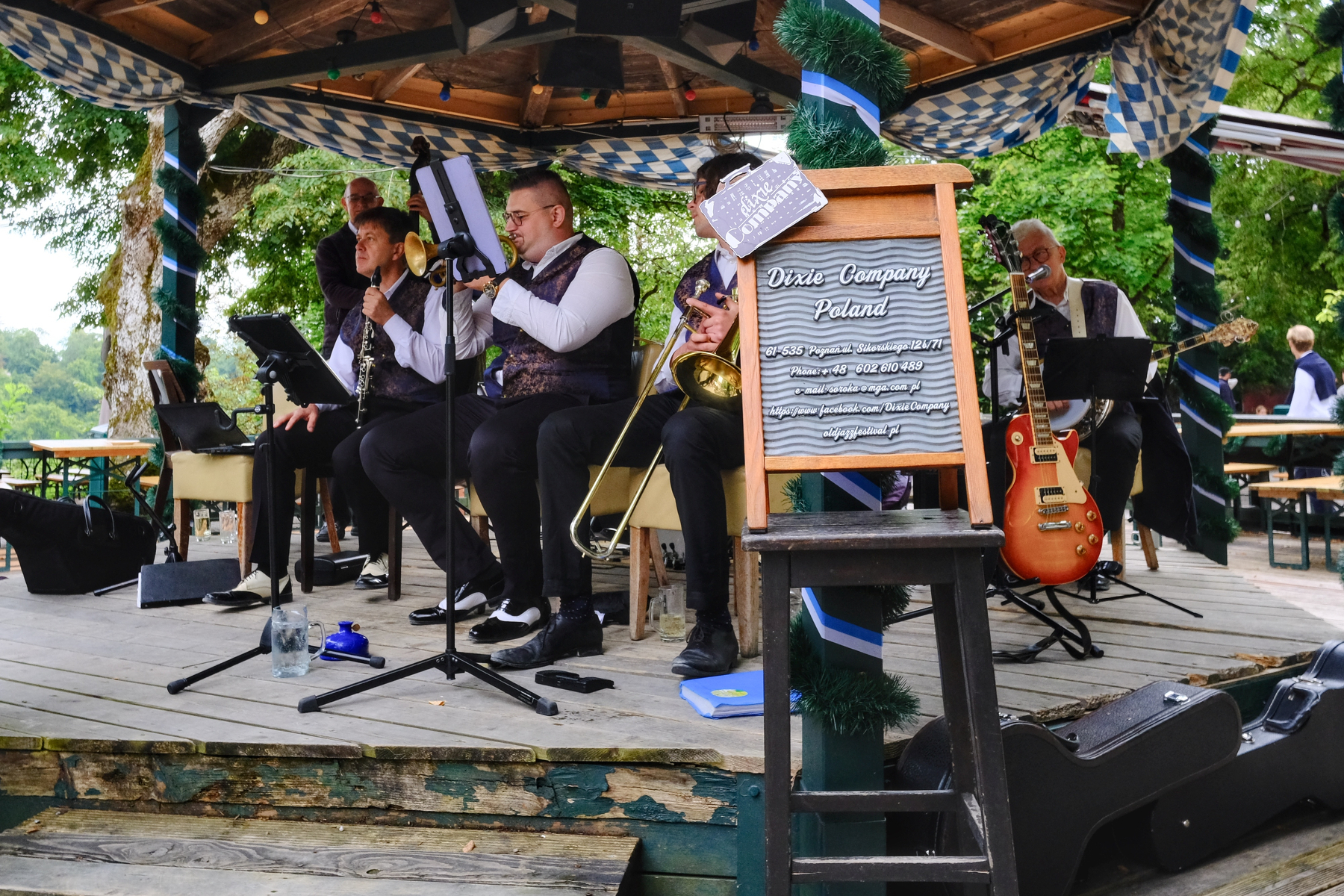 A group of musicians are playing under a large gazebo in the middle of a outdoor biergarten patio. You've got at least a clarinet, cornet, trombone, stand-up bass, guitar, and banjo. The sign in front says Dixie Company Poland